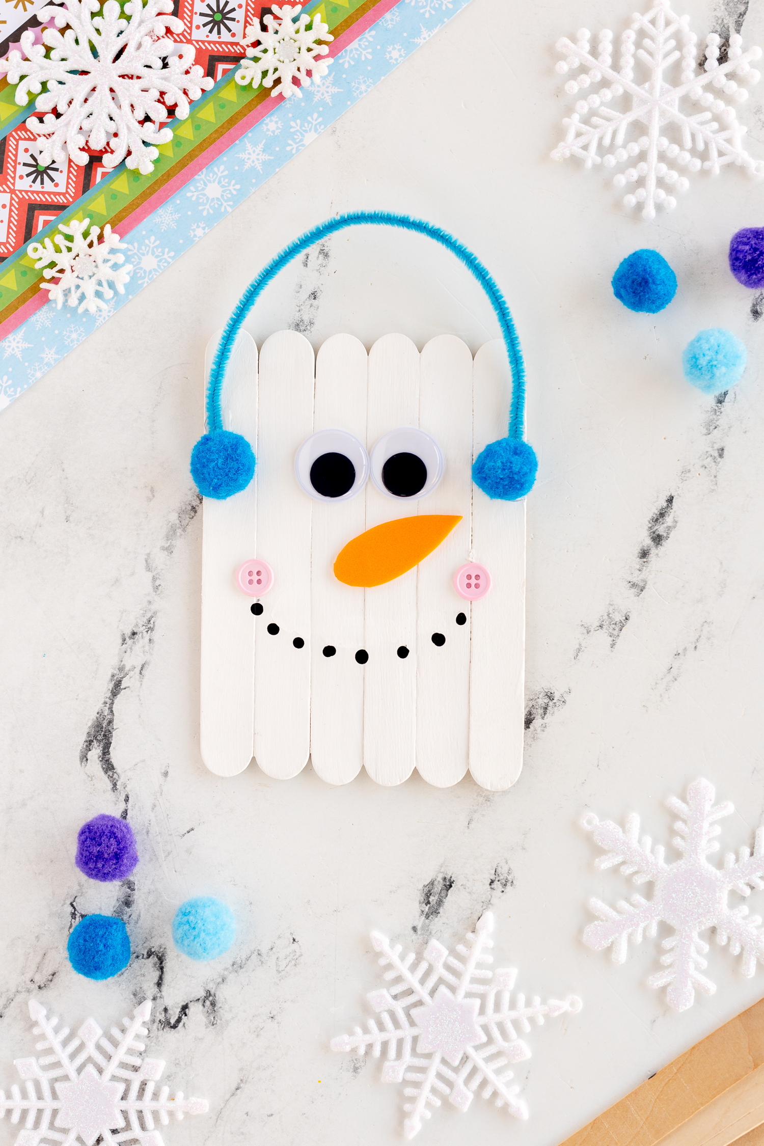 Popsicle Stick Snowman Craft with blue earmuffs made from pipe cleaner and pom poms