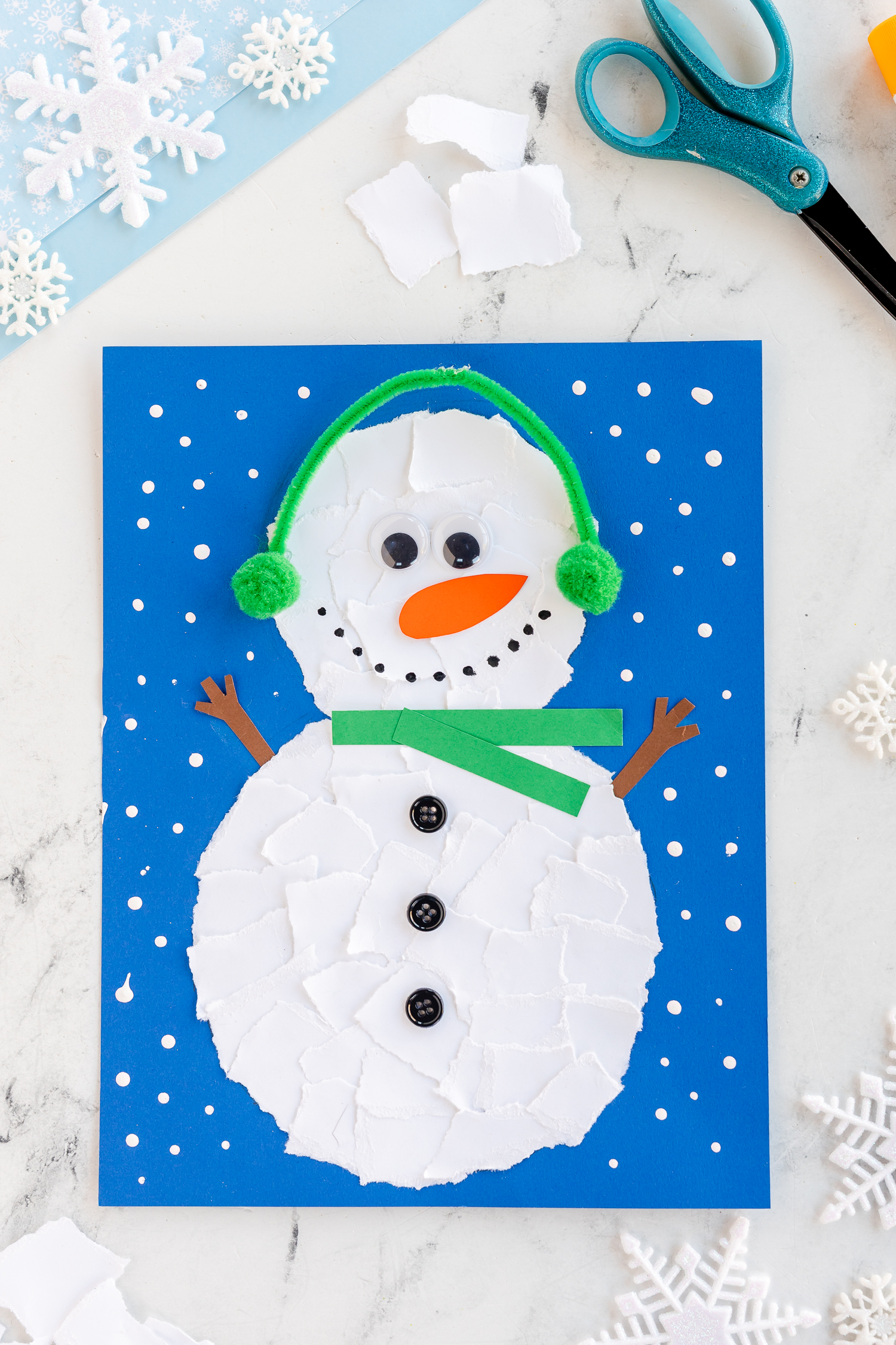 torn paper snowman craft on bright blue paper with green scarf and earmuffs
