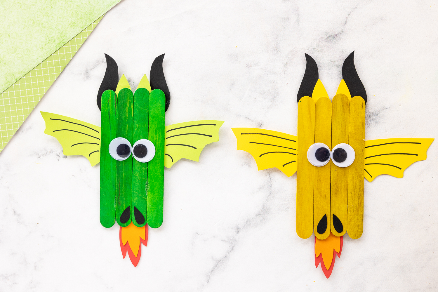 popsicle stick dragons on counter with green scrapbook paper nearby