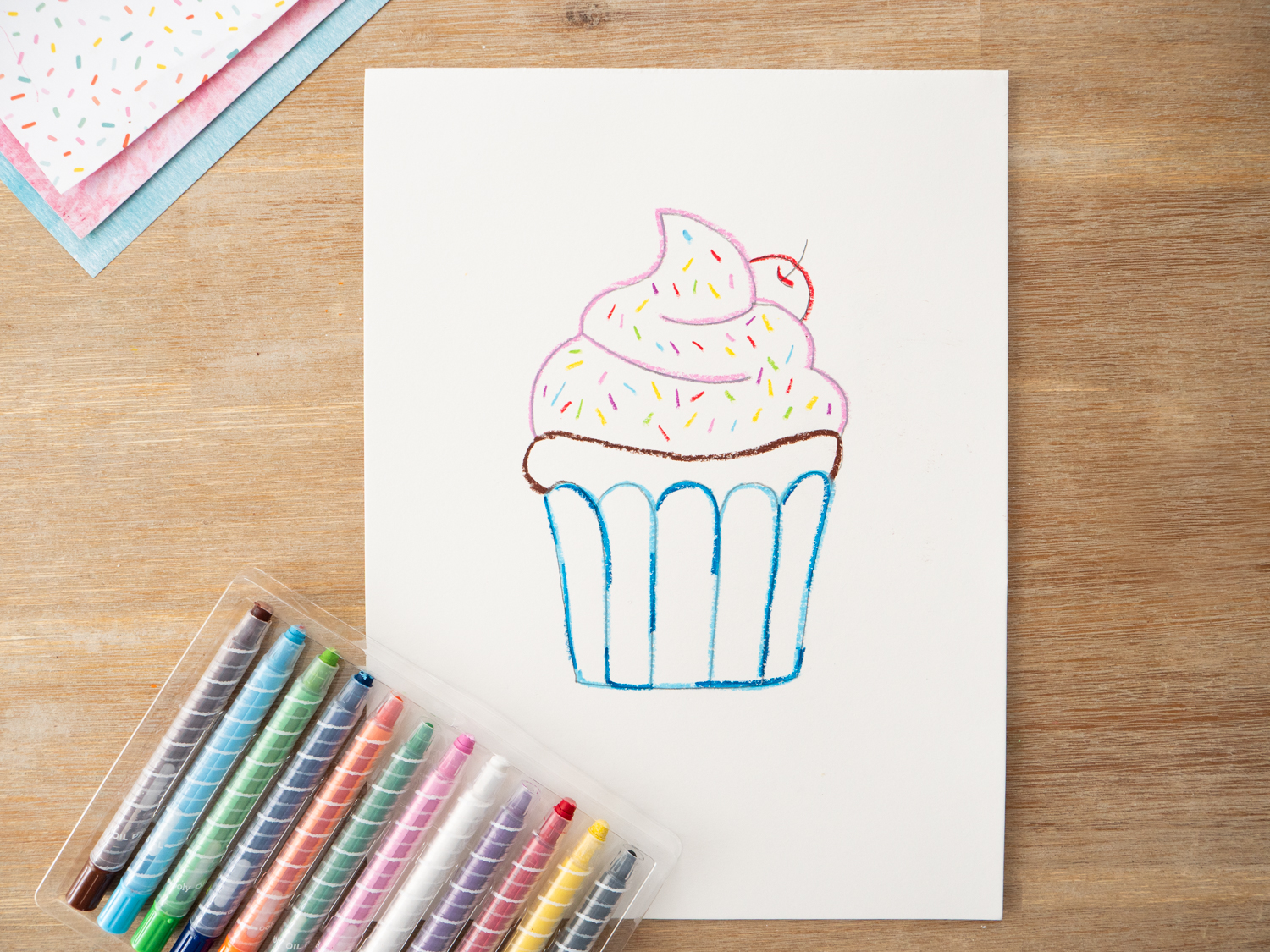 Cupcake art with oil pastel sprinkles, frosting details, and table line drawn before watercolor is added.