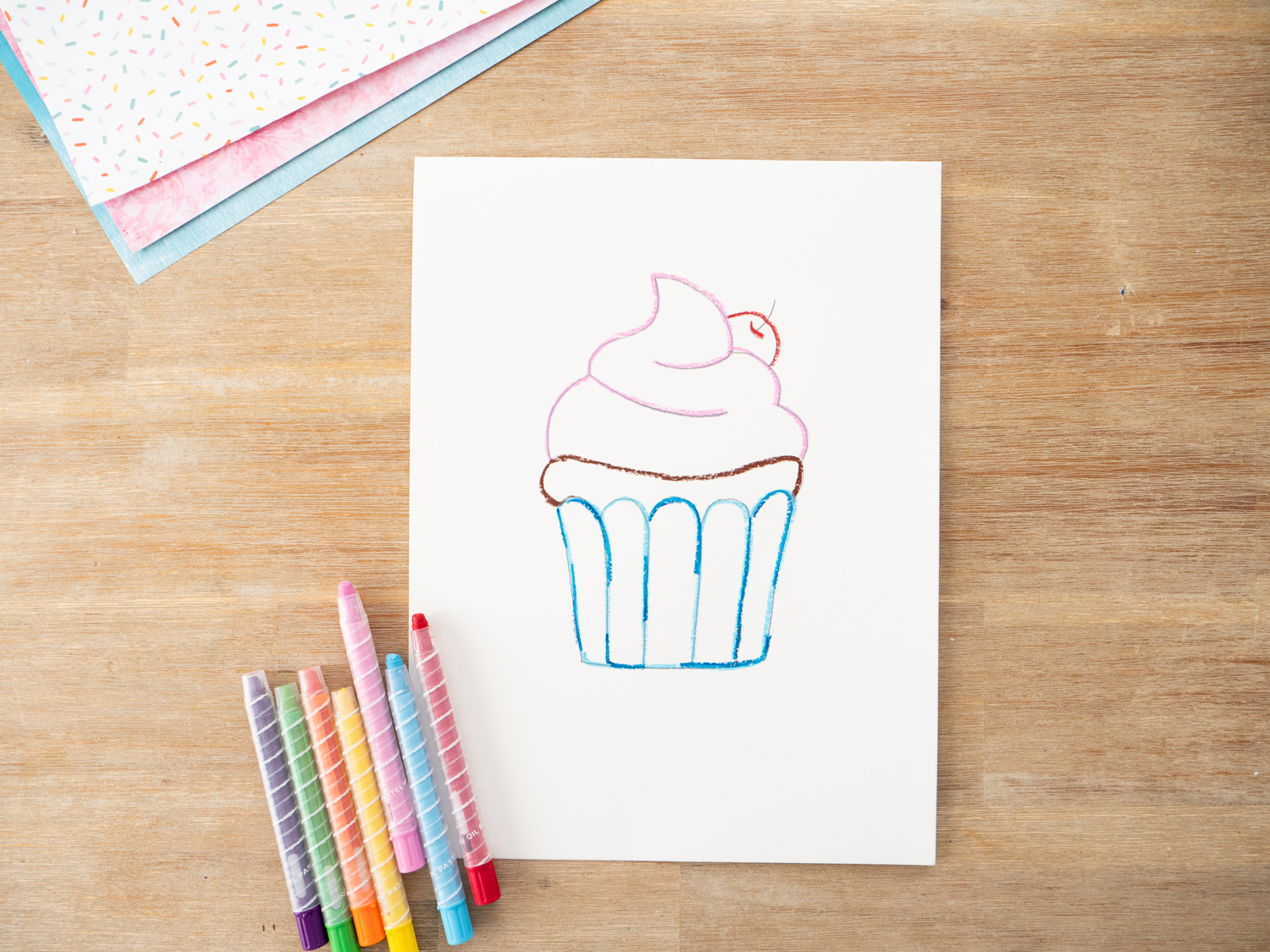 Cupcake art with oil pastel sprinkles, frosting details, and table line drawn before watercolor is added.