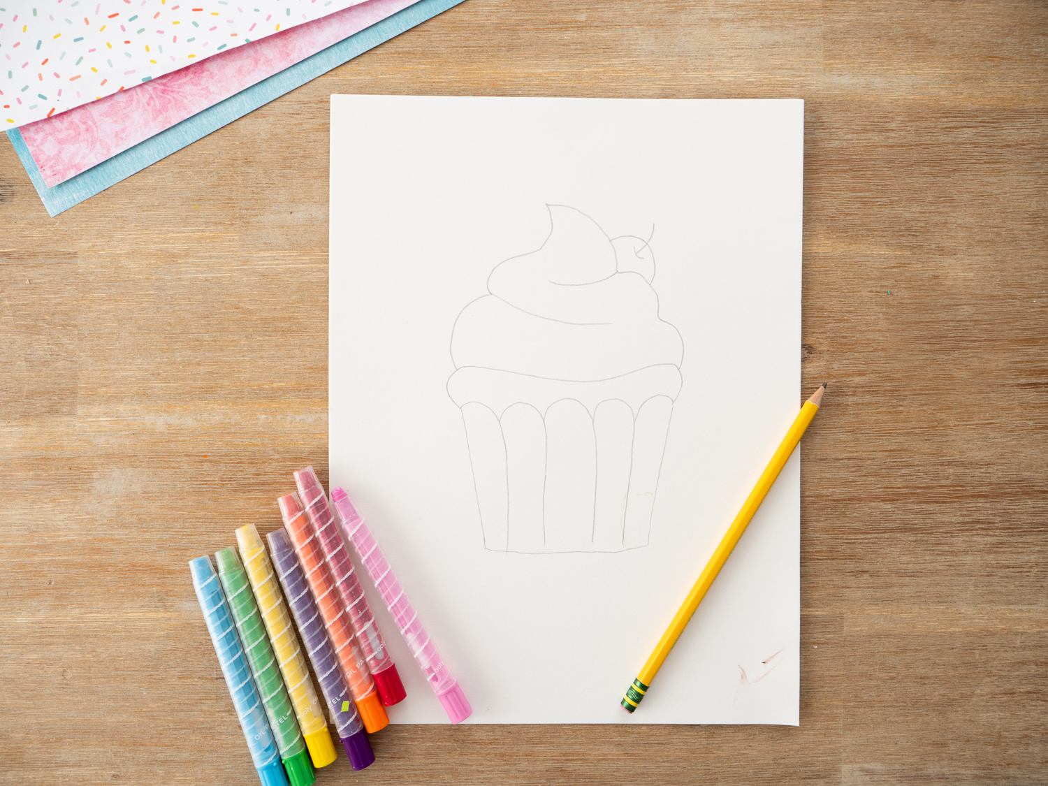 Child drawing cupcake outline with oil pastels on white watercolor paper before painting.