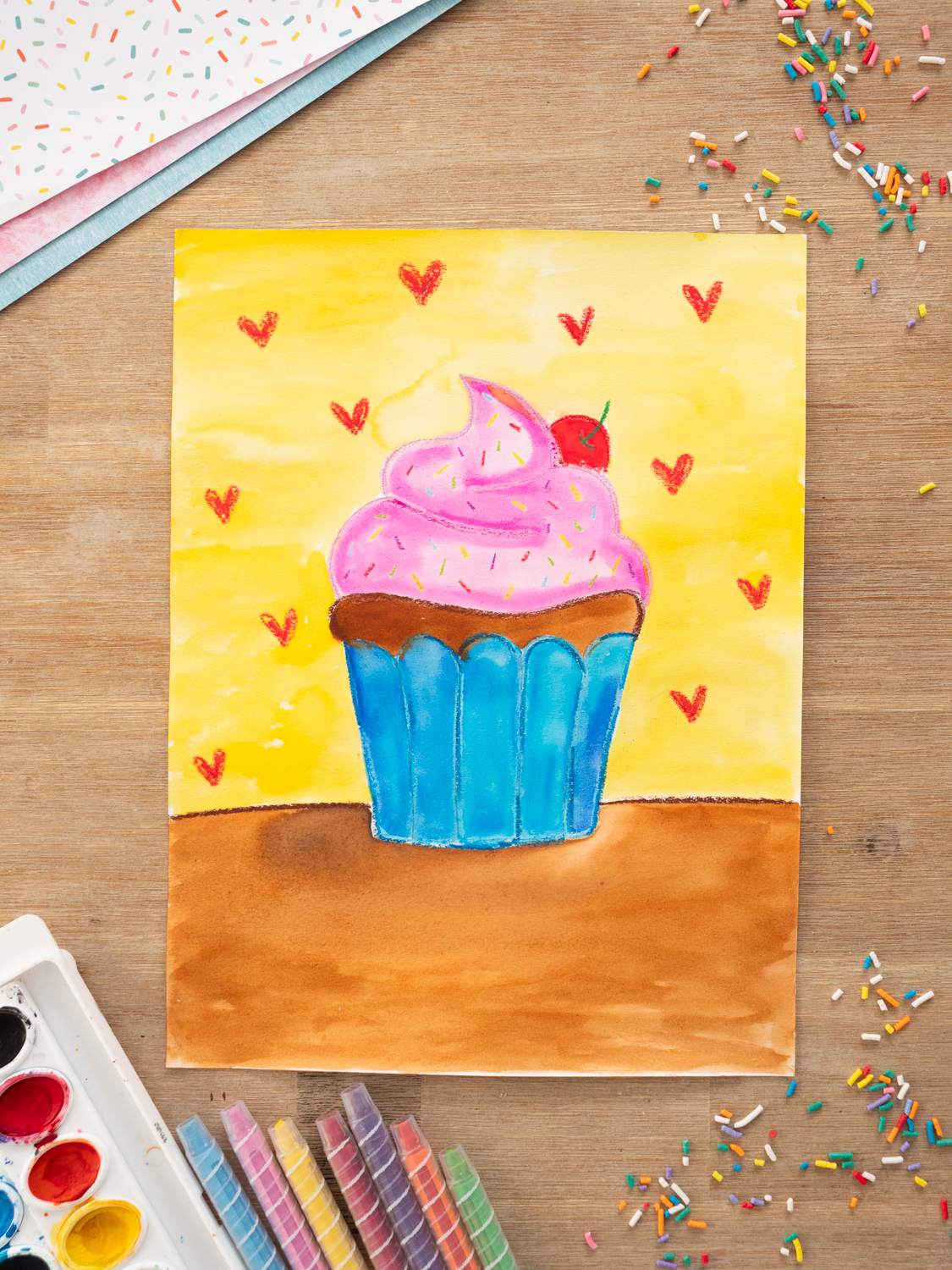 Watercolor cupcake art for kids featuring pink frosting, colorful sprinkles, blue cupcake wrapper, and yellow background using oil pastel resist technique.