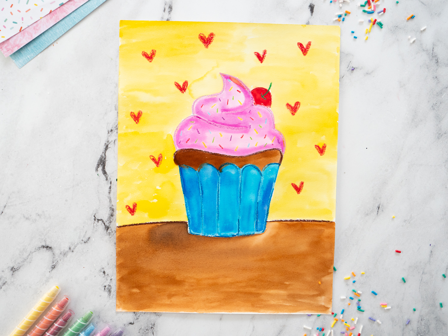 Close-up of watercolor cupcake art showing oil pastel resist lines and blended yellow background.